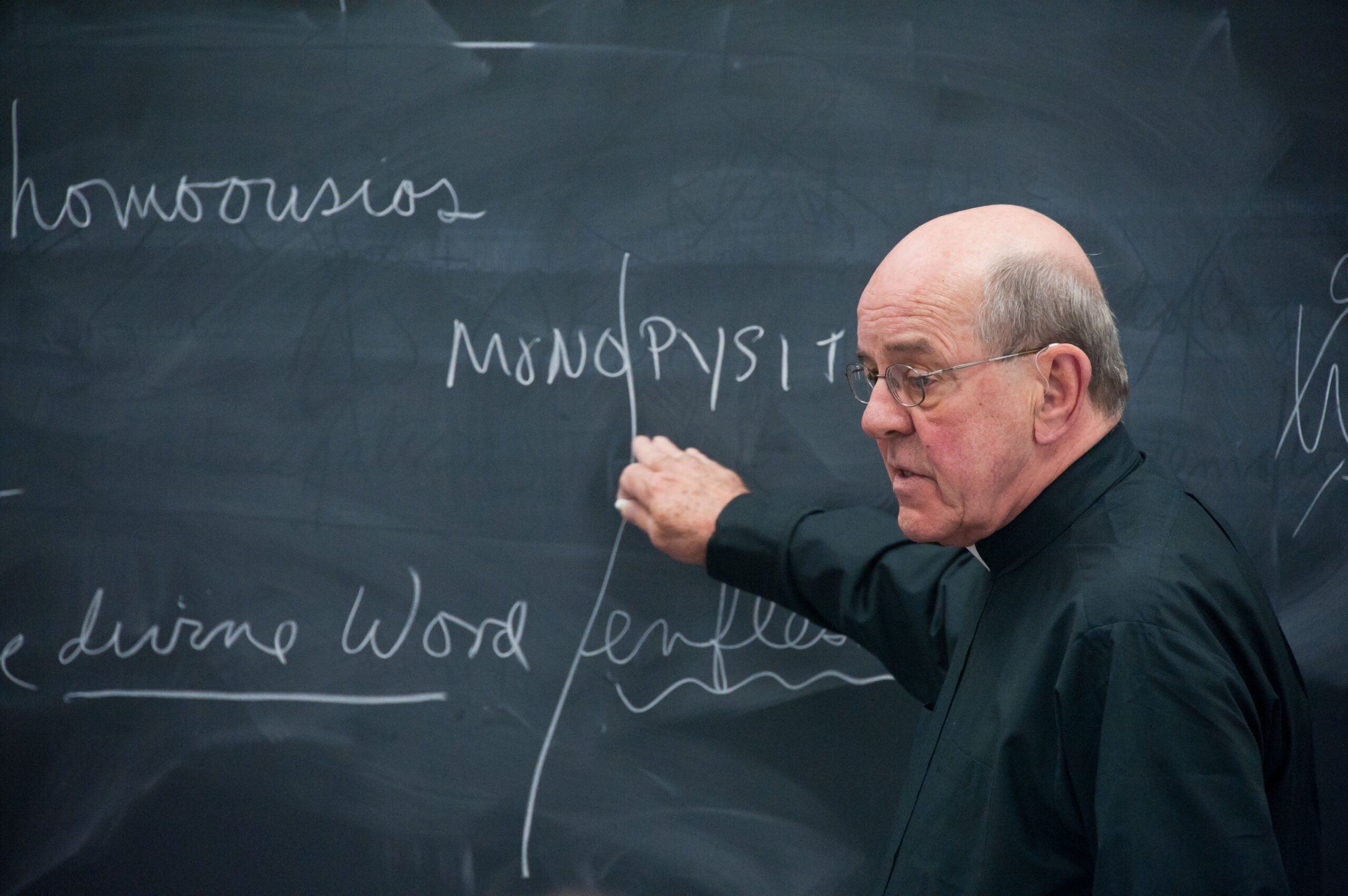Father Otto Hentz standing in front of and pointing at a chalk board with writing on it. H
