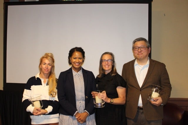 Four people stand in front of a white screen. From left to right: Meg Leta Jones is wearing a black and white striped shirt with a collar and is holding a glass award. Soyica Colbert is wearing a vertically striped dress with a dark blazer. Katherine Chandler is wearing a black dress and glasses and is also holding a glass award. Paul Ohm is wearing a white button up shirt and a brown blazer, he is also holding a glass award. 
