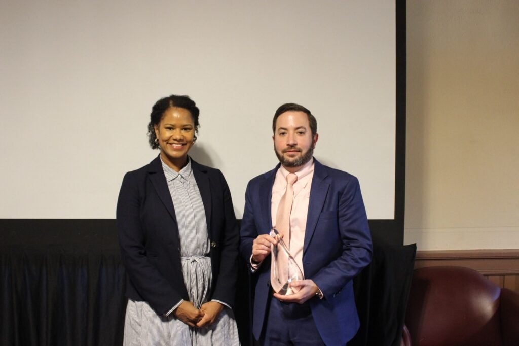 Two people stand in front of a blank screen. On the left, Soyica Colbert is wearing a vertically striped dress and dark blazer. On the right, Jeremy Fineman is wearing a navy suit with a pale pink shirt and tie. He is holding a glass award. 