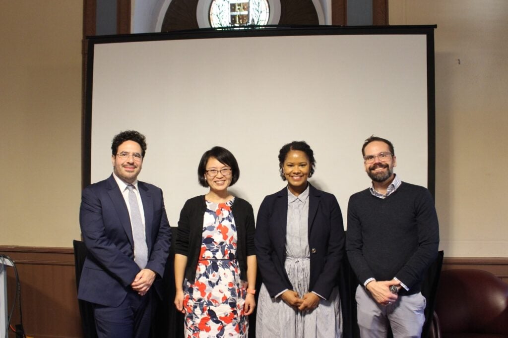 Four people stand in front of a blank screen. From right to left. Mike Amezcua is wearing a navy blue suit with a light gray tie. Britt He is wearing a patterned dress and a black cardigan. Soyica Colbert is wearing a vertically striped dress with a dark blazer. Shaun Brinsmade is wearing a dark sweater over a collared shirt. 