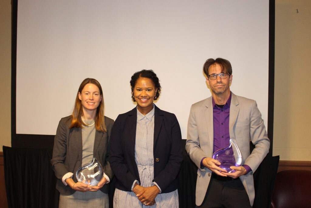 Three people stand in front of a blank screen. On the left Blythe Shepard is dressed in a gray dress and darker blazer, she is holding a glass award in the shape of a flame. In the middle is Soyica Colbert, she is wearing a dress with vertical stripes and a navy blue blazer. On the right is Ian Lyons wearing a purple shirt and a light gray blazer, he is also holding a glass award.